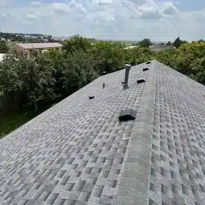 A gray shingled rooftop with several vents and pipes, overlooking trees and buildings.