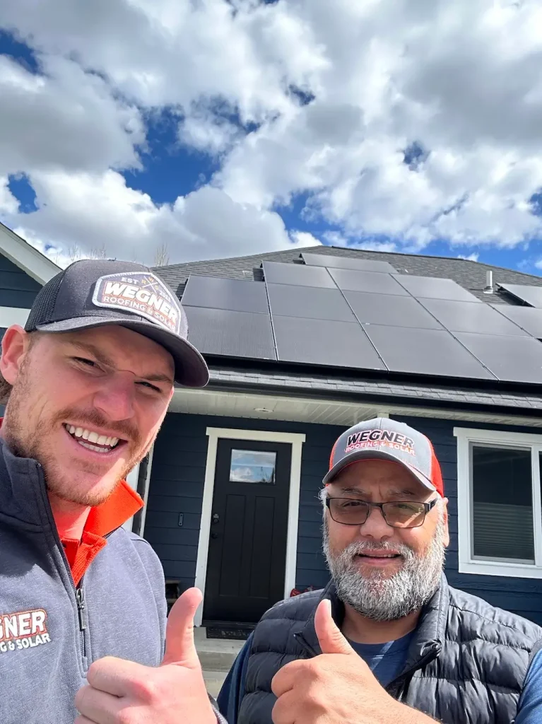 Two individuals gesturing thumbs up in front of a dark blue house with solar panels on the roof.