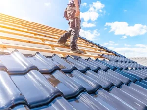A roofer stands on a framed roof, preparing to install glossy blue tiles under a bright sky with scattered clouds.