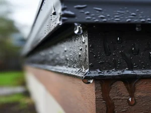 Close-up of water droplets clinging to a dark metal roof edge, with rainwater flowing down onto a wooden support below.
