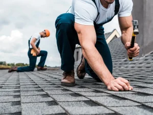 Two roofers work on a sloped roof, one kneeling with a hammer, securing shingles, while the other prepares materials nearby.
