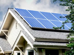 A house with a sloped roof featuring blue solar panels under a cloudy sky, surrounded by leafy trees.