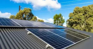 Solar panels installed on a gray metal roof with a clear blue sky and trees in the background.
