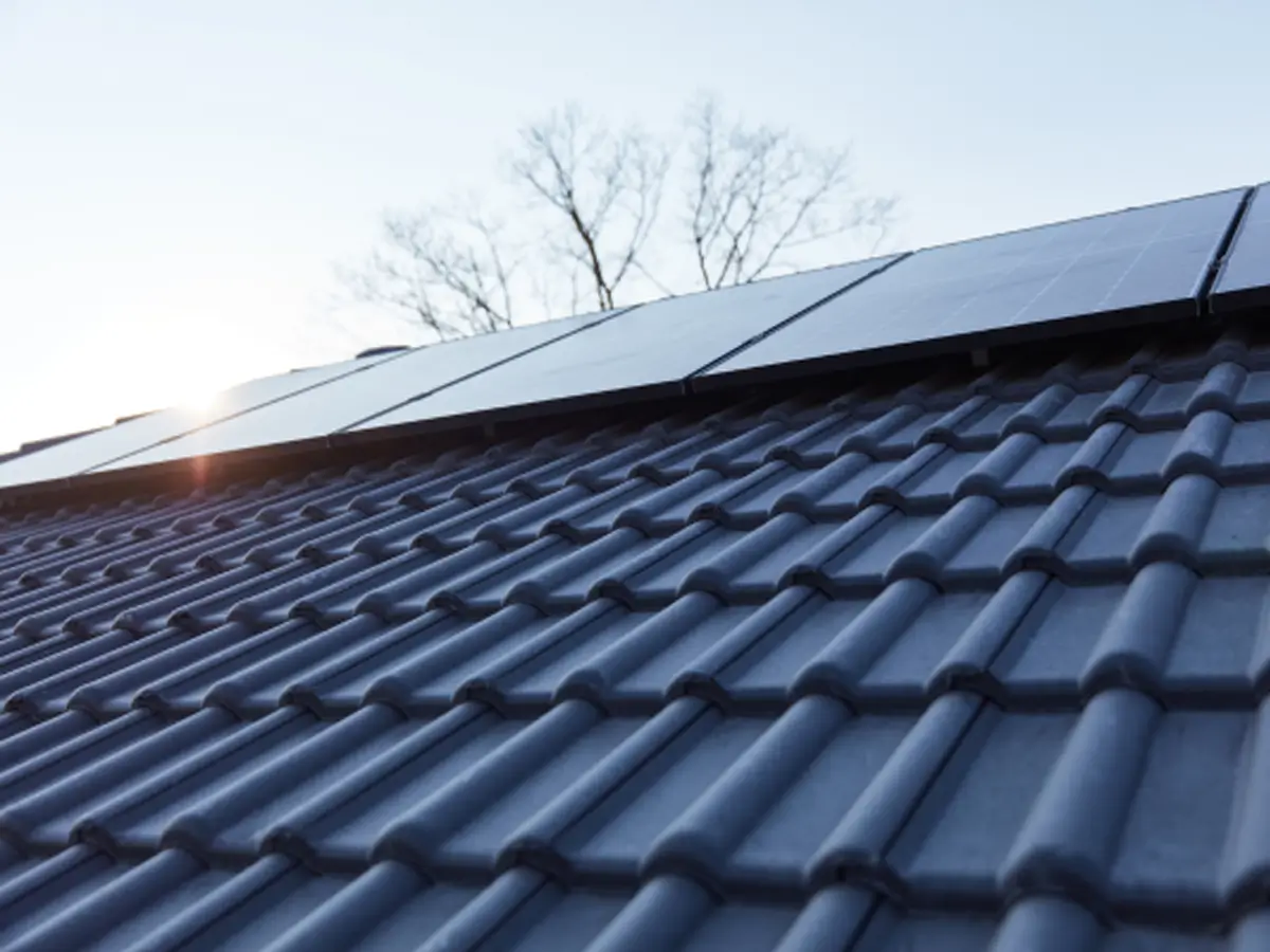 Close-up of a blue tiled roof with solar panels, sun rays shining through, and bare trees in the background against a clear sky.
