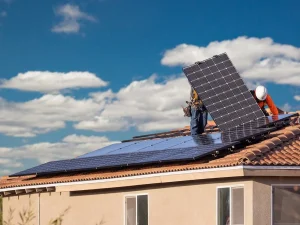 Workers install solar panels on a roof under a blue sky with fluffy clouds, promoting renewable energy solutions.