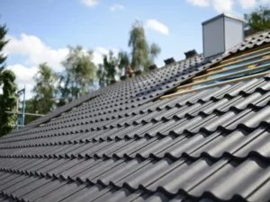 A close-up of a textured black roof with wavy tiles, under a blue sky with clouds and surrounded by green trees.