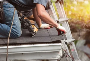 A roofer, equipped with a nail gun, fastens shingles on a roof while kneeling beside a ladder in a sunlit outdoor setting.